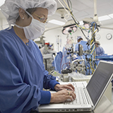 Infirmière portant une tenue chirurgicale complète tape sur un ordinateur pendant que des médecins opèrent en arrière-plan