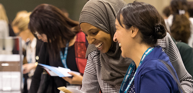 Deux femmes en train de rire et de parler devant l'affiche d'une université à la conférence Transformation Qualité de la santé 2015.