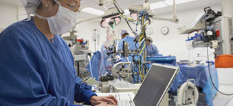 Nurse in full scrubs typing on a computer as doctors perform surgery in the background