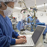 Nurse in full scrubs typing on a computer as doctors preform surgery in the background