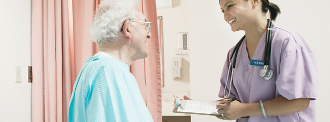 Female clinician stands bedside with an older male patient with a chart in her hand.