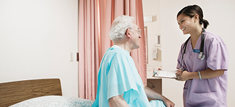 Female clinician stands bedside with an older male patient with a chart in her hand. 