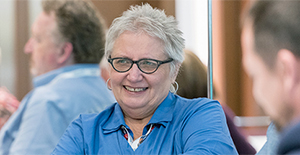 Gray-haired woman wearing blue shirt and glasses seated in a group smiles while interacting with others in a group