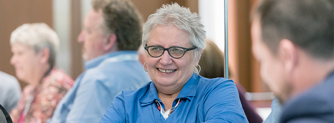 Gray-haired woman wearing blue shirt and glasses seated in a group smiles while interacting with others in a group
